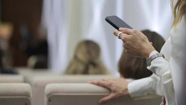 Woman holds phone while seated in auditorium. She checks device, ignoring surrounding audience. Her posture suggests focus or distraction. Scene captures modern multitasking during events