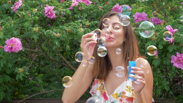 Young woman enjoy sunny day outdoor, blowing iridescent soap bubbles in a park with a beautiful pink rose bush in background. Representing carefree fun and happiness. Summer holidays in park