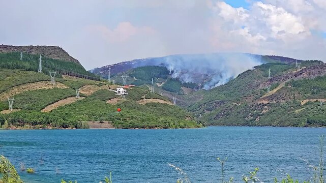 Firefighting helicopter collecting water from a lake with a bambi bucket to extinguish a devastating forest fire on a green mountain