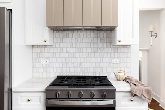 A kitchen range hood detail with a brown range hood, white cabinets and marble countertop, stainless steel appliances, and a marble tile backsplash.