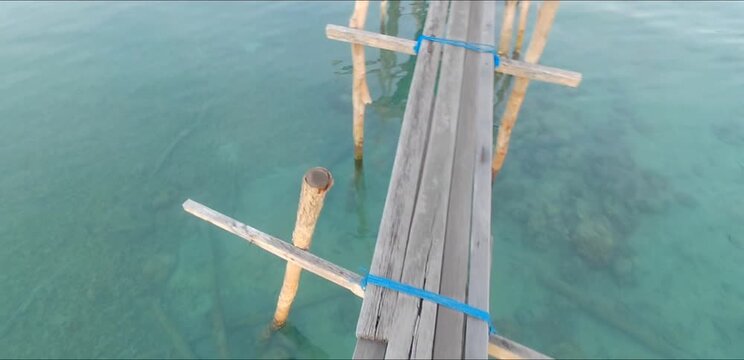 First-person POV tracking shot walking across a narrow, rustic wooden footbridge over clear turquoise tropical sea water.