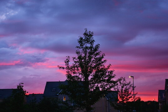 Hampton Water, Peterborough, England, UK - June 20 2026: Late sunset causing dramatic skyline with vibrant reds and blues behind the outline of rooftops and a tall tree