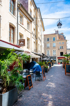 Sunny cobblestone square in Nancy, France, with an outdoor restaurant terrace featuring palm trees, chalkboard menus, rattan chairs, and a vintage hanging lantern, with classic French stone buildings 