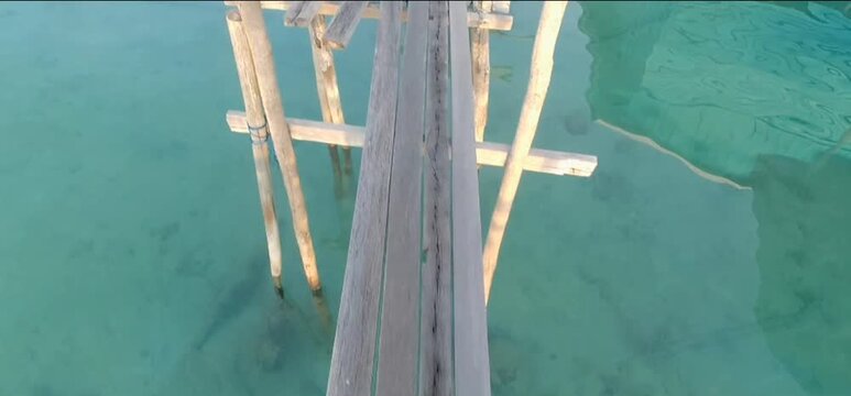 First-person POV tracking shot walking across a narrow, rustic wooden footbridge over clear turquoise tropical sea water.