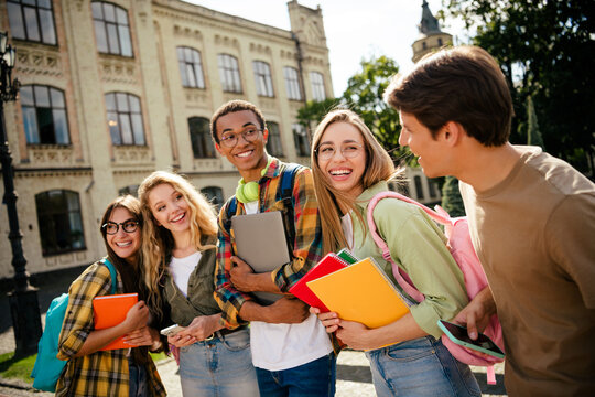 Group of smiling university students and friends on campus carrying notebooks and backpacks, diverse young people laughing and talking outdoors by historic campus buildings