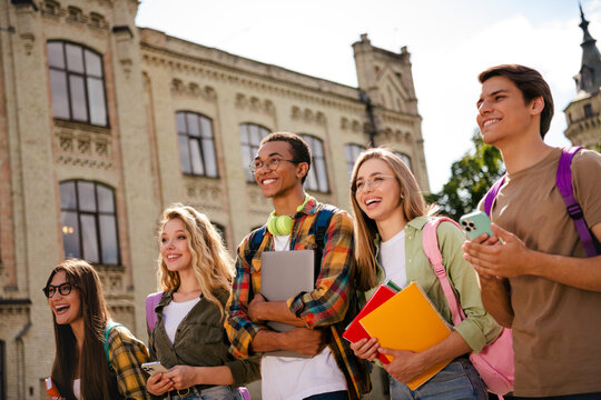 Smiling diverse college students walking outside historic campus with backpacks, notebooks and laptop enjoying friendship and campus life