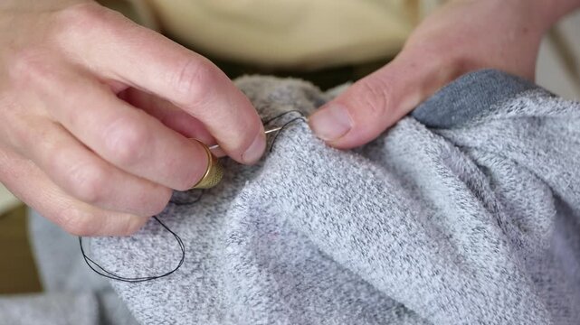 Close-up of hands sewing clothes with a thimble. Careful female hands using a needle, black thread, and a thimble to sew a hole in a grey sweater. A detailed view of the traditional mending process