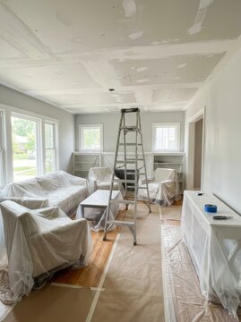 Interior room renovation with furniture covered in plastic sheeting. A ladder stands in the middle of a home undergoing ceiling repair and preparation for painting. Residential DIY.