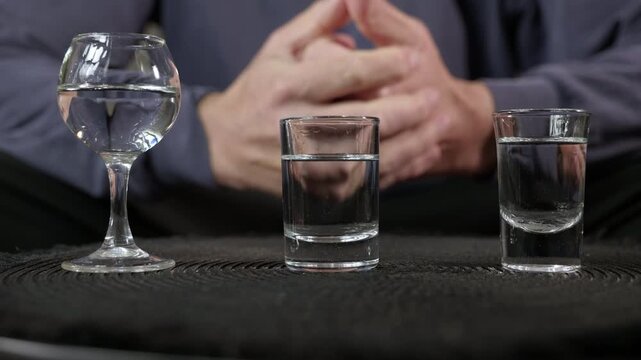 Stressed man with glasses of alcohol. Nervous man wringing his hands behind three glasses filled with clear alcohol, expressing anxiety, stress, and hesitation before facing an addiction problem