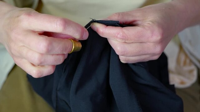 Seamstress hands sewing black fabric with needle and thread. Close-up of a tailor's hands carefully sewing a hem on a piece of black cloth using a needle, thread, and a thimble, demonstrating mending