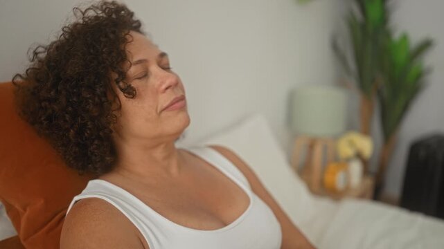 Woman reclining on bed in a building wearing a white tank top, chest visible, eyes closed and mouth slightly open while leaning back on a rust pillow; calm rest.