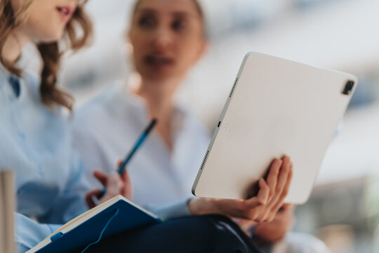 Two business colleagues review a tablet while taking notes and discussing ideas. The women collaborate in an informal meeting, using digital technology and a notebook.
