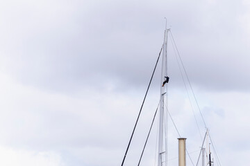Obraz premium Distant view of sailor climbing high sailboat mast for maintenance under overcast cloudy sky