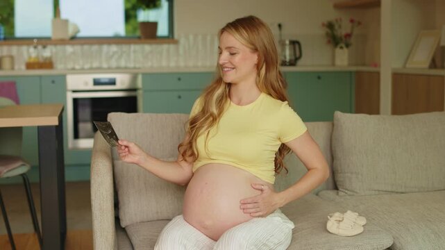 Expectant mother smiling at prenatal sonogram image while softly stroking her belly during peaceful moment at home, 4K, cinematic footage