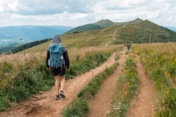 Fototapeta premium Back View of a Female Hiker with a Backpack and Hooded Sweatshirt Walking on a Mountain Ridge Dirt Trail Toward Distant Green Peaks