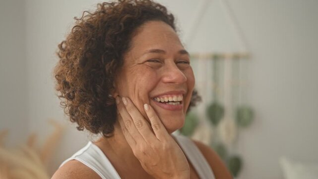 Woman touching cheek with hand while smiling and laughing beside a macrame wall hanging and plants in studio; joy wellness comfort.