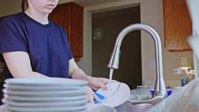 Water flows from the faucet as a person cleans a dish in a kitchen. A stack of plates sits on the counter beside the sink during the day.