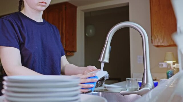 A person stands at a kitchen sink washing dishes with a sponge in hand. Plates are stacked beside the sink. Water flows from the tap as the task is being done.