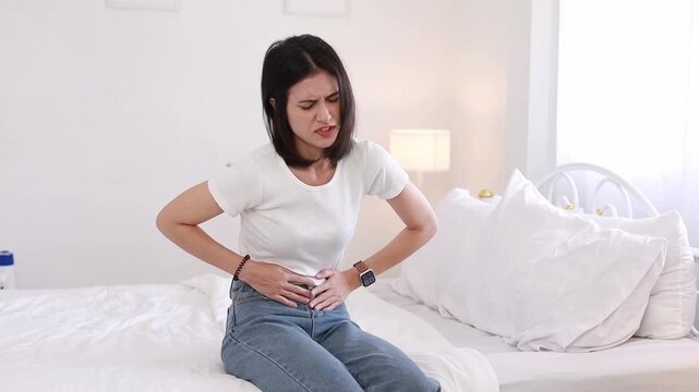 A young woman sat clutching her stomach due to food poisoning, experiencing abdominal pain and intestinal problems such as gastritis or diarrhea while at home.