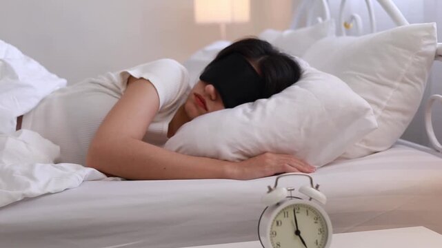 A sleepy young woman with an eye mask is turning off the alarm clock on her bedside table. She presses the alarm button, waking up to the sound of the alarm while she was sleeping soundly.