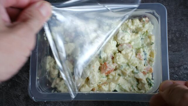 Person hands carefully peeling back plastic film from rectangular container filled with fresh, ready to eat potato salad on dark, textured surface, preparing convenience meal. Semi finished products