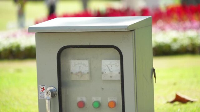 An electrical control box featuring voltage and ammeter gauges at the top, multiple red, green, orange, and white push buttons, and a metal handle on the left side, situated outdoors on grass in dayli
