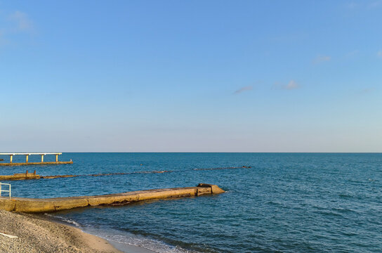 A seashore at sunset with a partially submerged breakwater against a blue sky; the sun is setting below the horizon.