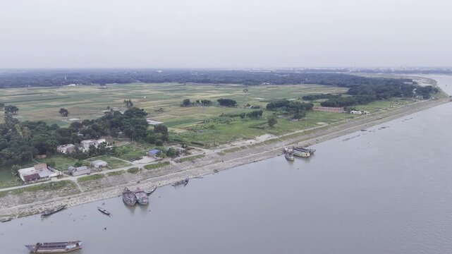 Aerial view moving over a wide river with traditional boats, a riverside settlement, green farmland, and a rural landscape in Bangladesh.