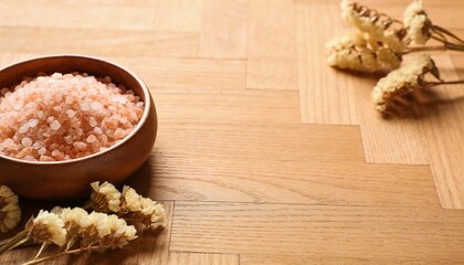Fototapeta premium A wooden bowl filled with pink sea salt rests on a textured wooden surface, accompanied by dried flowers