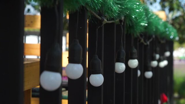 A row of white bulb lights hangs from a black metal railing decorated with green garland, photographed in soft daylight with a shallow depth of field.