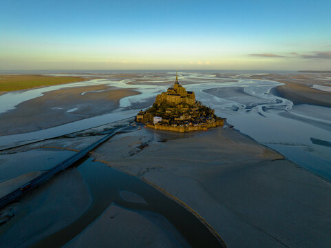 Aerial view of the historic Mont Saint-Michel abbey surrounded by vast tidal flats at low tide during golden hour in Mont Saint-Michel, Normandy, France.