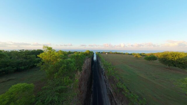 Smooth Cinematic Aerial Flight Above Deep Road Cut Through Coastal Cliffs At Pantai Tanah Barak, Wide Angle Sunrise View Toward Ocean Horizon And Green Landscape, Journey And Discovery
