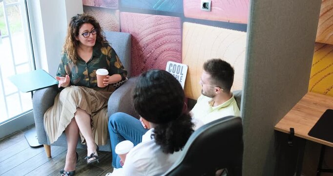 Three diverse colleagues sitting together in a colorful modern office lounge, having a casual conversation during a coffee break.