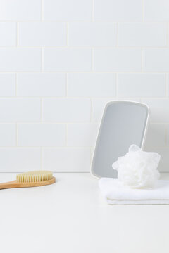 Bathroom product mockup background with white subway tile wall. Empty vanity countertop with mirror, towel, bath puff, and wooden brush for skincare cosmetic display.