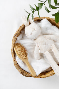Flat lay of baby care product mockup background with plush bunny toy, wooden brush, and white towel in wicker basket. Top view with green leaves and copy space for organic newborn skincare brand.