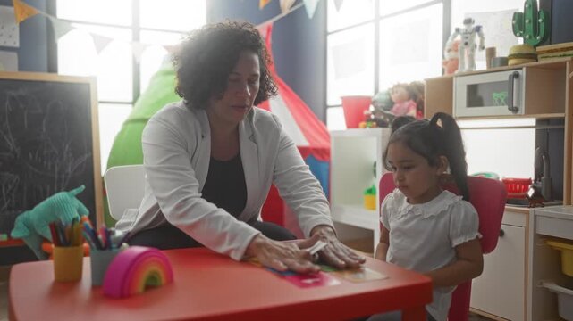 Woman teacher places hands on colorful numbered cards on a small red table while little girl watches attentively in a preschool building; early learning calm.