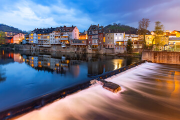 Fototapeta premium Illuminated houses along the Lenne River in Hagen-Hohenlimburg (Germany) at twilight. An atmospheric panoramic long-exposure shot featuring a cascade. The buildings stand directly on the riverbank and