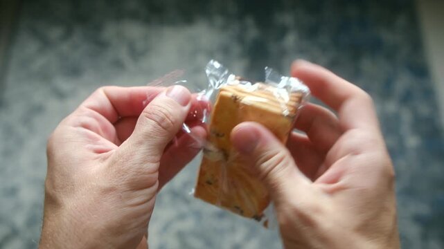 Man hands unwrapping small, transparent plastic package of raisin crackers, getting ready for quick snack during break over blurred, textured background. Person hands opening pack of raisin crackers