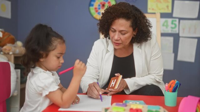 Teacher woman holds letter flashcard h and purple pen while smiling and leaning toward girl at red table in school; learning warmth patience.