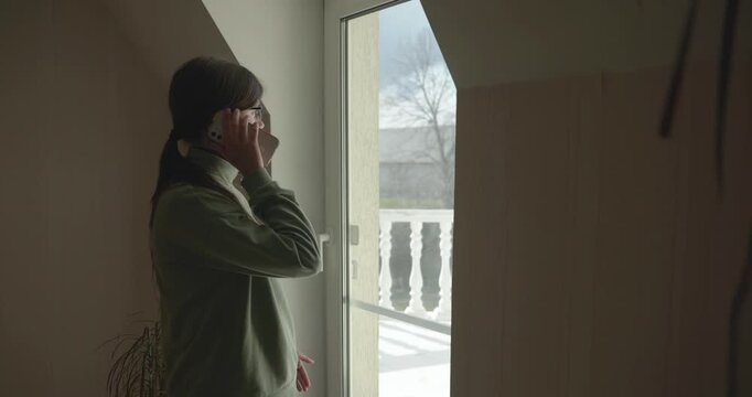 Thoughtful Girl Talks on Phone Looking out of Window. 4K 10-bit