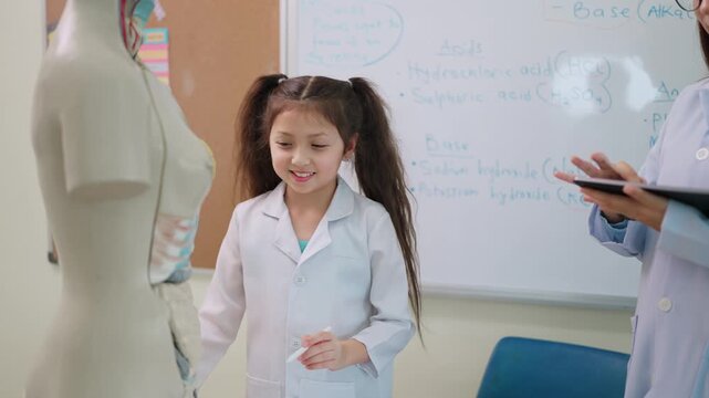 Young girl in lab coat explores human anatomy. She points at a mannequin's chest with curiosity. Teacher stands nearby, guiding her learning. Bright classroom with whiteboard and science posters