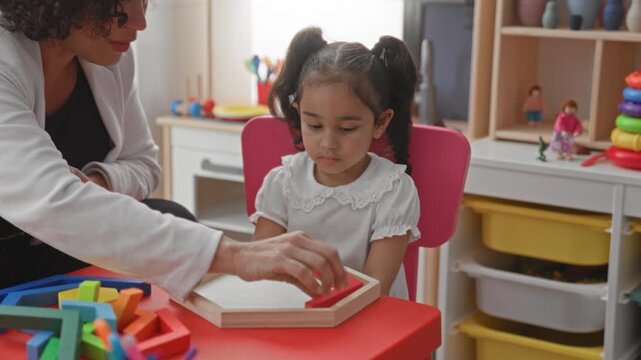 Teacher woman guides little girl placing yellow block into wooden tray on red table in building; early learning concentration.