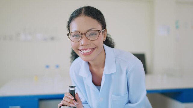 Young scientist smiles while examining samples through microscope. Lab setting includes beakers, laptops, and organized shelves. Professional, confident, and curious demeanor radiates in bright lab