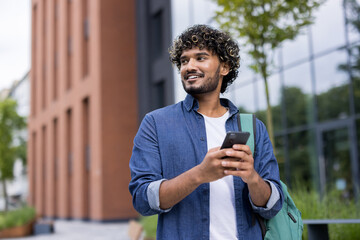 Obraz premium Young man with curly hair and backpack walking on a college campus while looking away and smiling, using his mobile phone for communication and connection