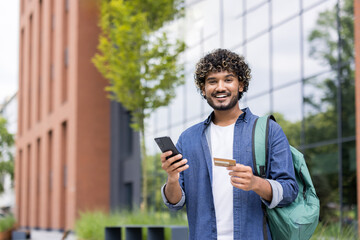 Fototapeta premium Smiling student with curly hair holding a smartphone and credit card for online transactions, shopping, and banking outdoors near a modern building