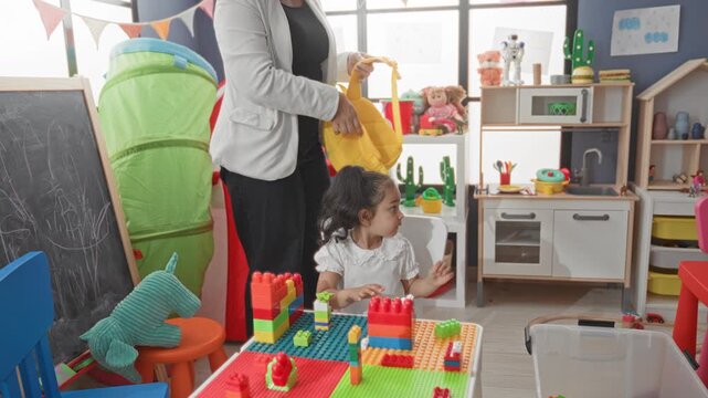 Woman teacher helps little girl put on a yellow backpack, adjusting straps while she sits at a toy table in a preschool classroom building; reassurance readiness.