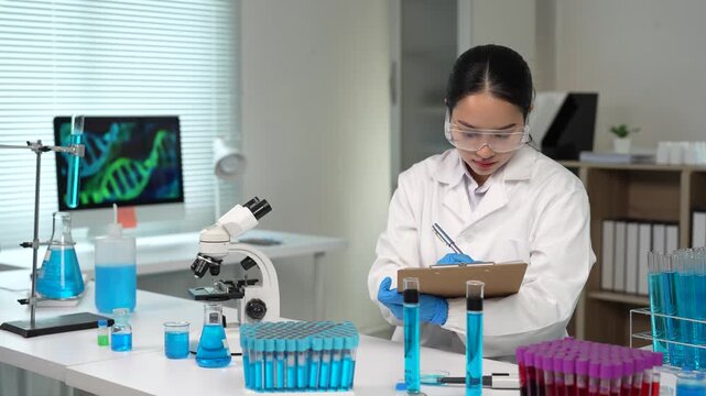 Asian scientist analyzing liquid samples in a genetics laboratory while a DNA strand is visible on a computer screen in the background, a concept of medical research