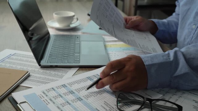 man Hands review financial reports with a calculator and pen beside a laptop, checking figures on printed documents for accounting analysis, budgeting records, and business finance workflow.