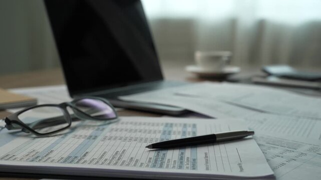 Pen and eyeglasses rest on printed financial documents beside a laptop on a desk for accounting report review, paperwork analysis, budget planning, and business finance content in an office workspace.