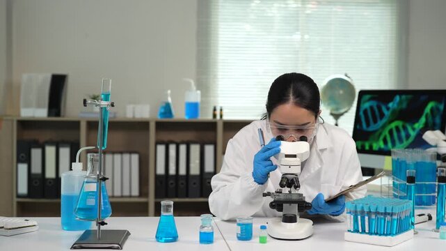 Asian scientist looking through a microscope and writing notes on a clipboard in a research laboratory conducting a scientific experiment with blue liquid and test tube